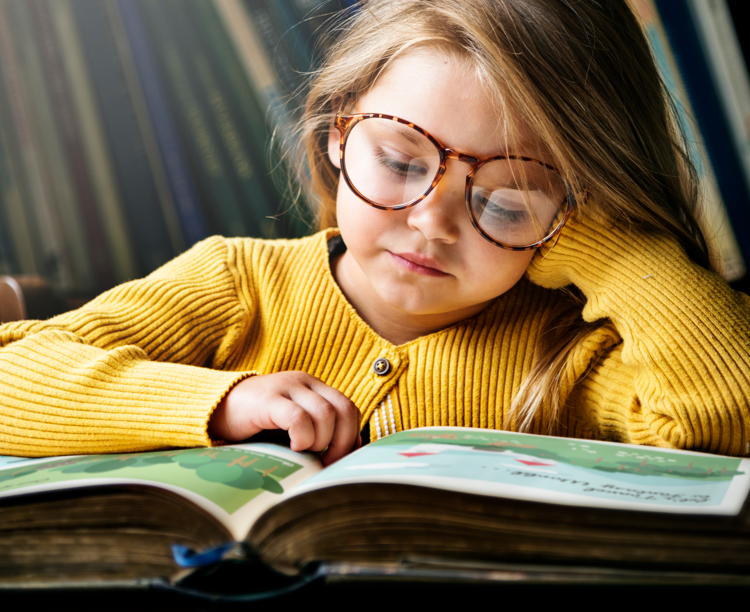 Niña leyendo un libro con lentes puestos.